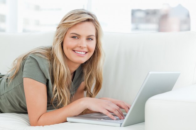 Mujer que miente en el sofá con la computadora portátil delante de ella, sonriendo