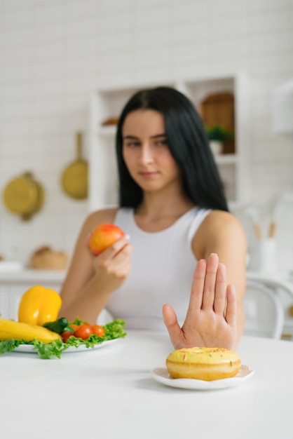 Mujer que rechaza la comida dulce y prefiere una nutrición saludable, tábanos y frutas sentadas en la mesa de la cocina