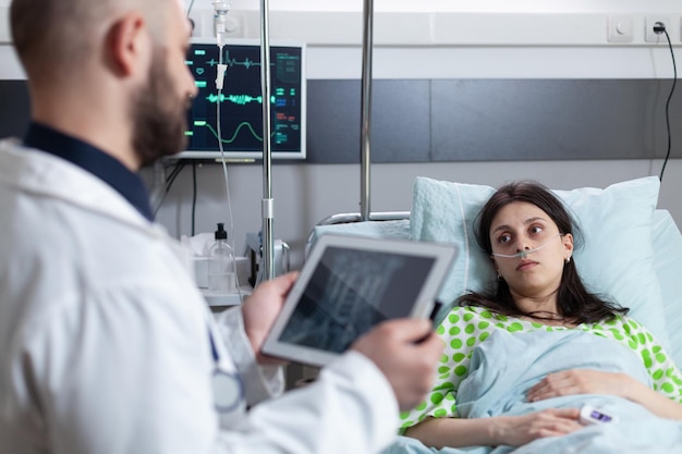 Mujer que recibe oxígeno y tiene controles vitales después de una intervención quirúrgica mirando al médico con una resonancia magnética en una tableta digital. Paciente en recuperación escuchando los resultados de imágenes de lectura médica.