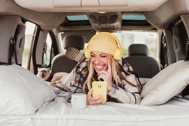Una mujer que se ríe colocando una mini autocaravana interior usando un teléfono móvil y escuchando la música con auriculares amarillos Gente nómada digital Travel vanlife