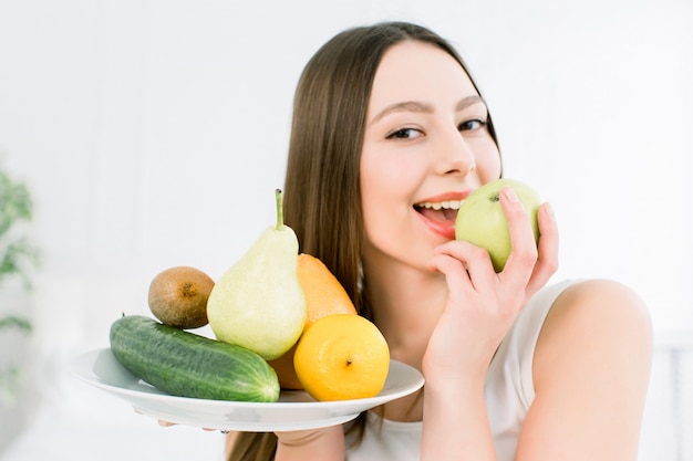 La mujer que sostenía el surtido de las frutas sirvió en la placa blanca contra fondo ligero. Retrato del primer de la mujer joven alegre con los dientes perfectos que muerde la manzana verde. Concepto de belleza saludable