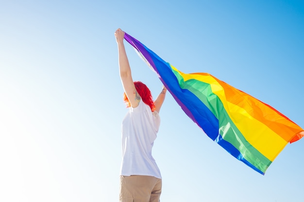 Mujer que sostiene la bandera gay del arco iris en fondo del cielo azul. Concepto de felicidad, libertad y amor para parejas del mismo sexo.