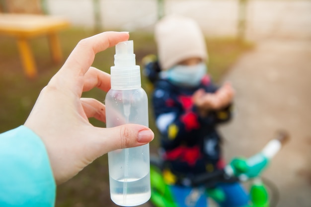 Mujer que sostiene un desinfectante en su primer de las manos. Desinfección de las manos de un niño caminando por la calle. Niño en una máscara médica en bicicleta, sostiene sus manos para desinfección