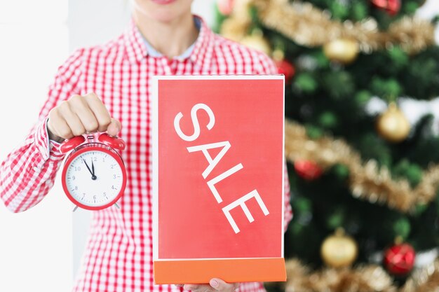 Mujer que sostiene la placa roja del reloj y de la venta de la alarma cerca del primer del árbol de año nuevo. Concepto de descuentos de promociones de año nuevo