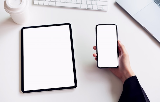 Mujer que sostiene smartphone y la tableta de la pantalla en blanco en la tabla.