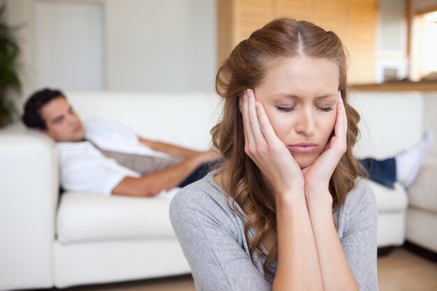 Mujer que tiene dolor de cabeza mientras que el hombre está acostado en el sofá detrás de ella