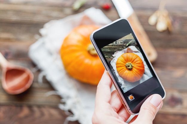 Mujer que tira la calabaza anaranjada fresca en la tabla de madera usando smartphone.