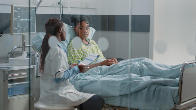 Mujer que trabaja como médico para tratar a un paciente joven con enfermedad en la cama. Médico general haciendo consulta médica con una persona enferma en la sala del hospital y tomando notas. Tratamiento sanitario