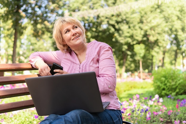 Una mujer que trabaja con una computadora portátil en un banco del parque. Freelancer mujer de mediana edad. Estudia correo electrónico, redes sociales.