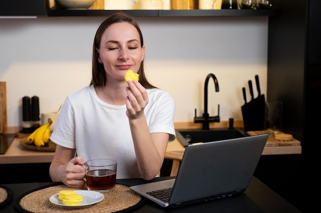 Mujer que trabaja en una computadora portátil en una oficina en casa una niña come pastel y bebe té en la cocina