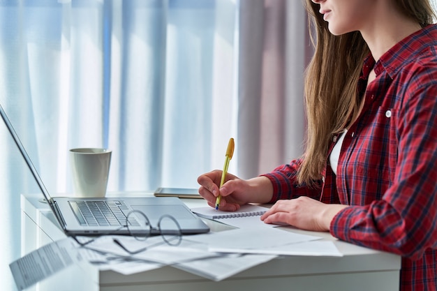 Mujer que trabaja remotamente en la computadora portátil y anota la información de datos importantes en el cuaderno. Estudiante durante la educación a distancia y cursos en línea que aprenden en casa