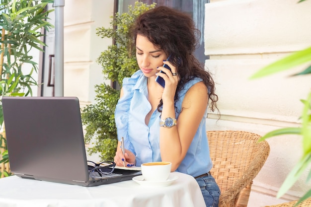 Mujer que trabaja remotamente con la computadora portátil y el teléfono en la cafetería. Feliz empresaria llamando por teléfono móvil y teniendo. Hermosa morena con notebook en café. Lanza libre