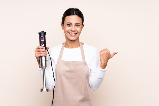 Mujer que usa la batidora de mano sobre una pared aislada que apunta hacia un lado para presentar un producto