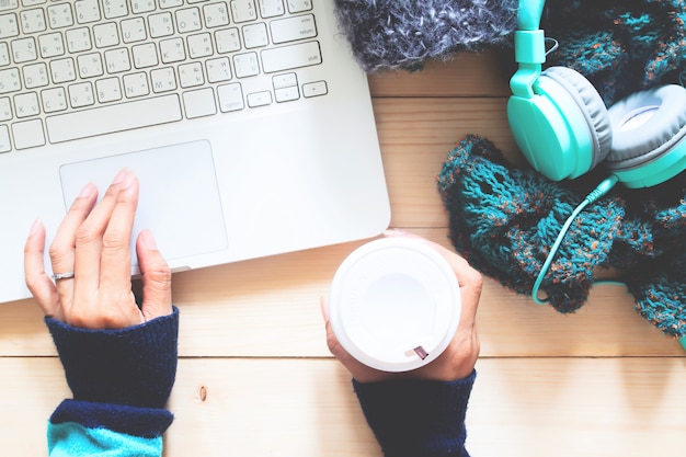 Mujer que usa la computadora portátil y bebiendo café, auriculares y bufanda en la mesa. Con otoño o invierno