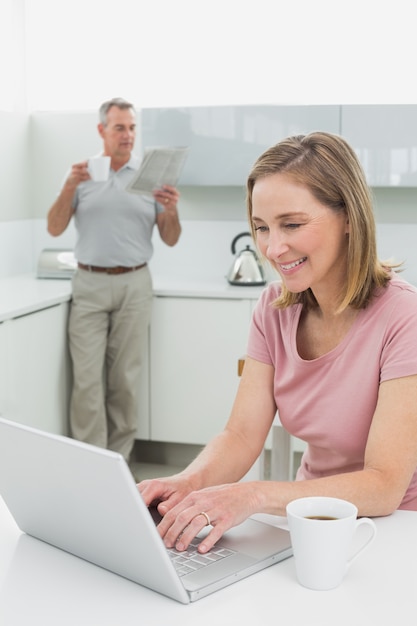 Mujer que usa la computadora portátil mientras que el hombre con la taza de café y el periódico en cocina