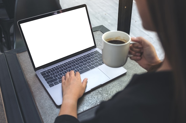 Una mujer que usa una computadora portátil con una pantalla de escritorio blanca en blanco mientras bebe café caliente en un café