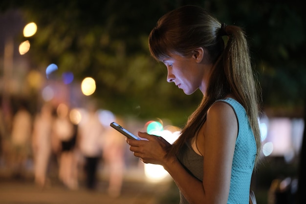 Foto mujer que usa el dedo acusador del teléfono en la pantalla del teléfono inteligente en el fondo de las luces bokeh en la iluminación atmosférica de la ciudad de noche en la calle desenfocada de noche. estilo de vida online con conexión a internet