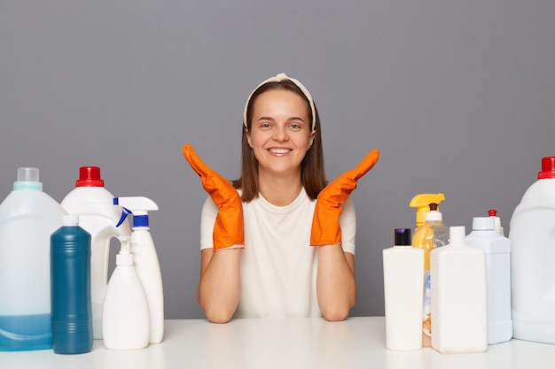 Una mujer que usa guantes protectores en la cabeza y una camiseta informal extiende las manos en el lugar de trabajo con muchos detergentes anuncia buenos suministros de limpieza para limpiar aislados sobre fondo gris