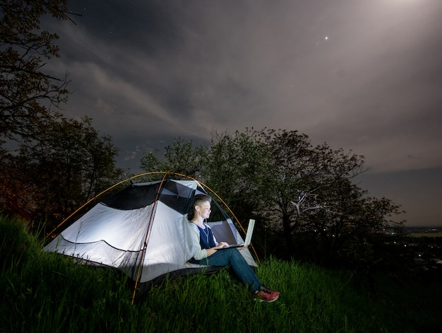 Mujer que usa su computadora portátil con las auriculares en el camping en la noche.