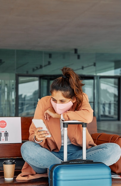 Foto mujer que usa su teléfono inteligente en el aeropuerto mientras se apoya en su equipaje durante la pandemia