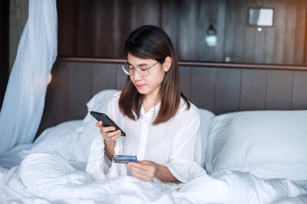 Mujer que usa un teléfono inteligente móvil y una tarjeta de crédito para comprar en línea mientras hace un pedido en la cama por la mañana en casa. tecnología, comercio electrónico, pago en línea de banca digital y concepto de vida en apartamentos