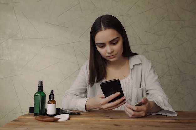 Mujer que usa un teléfono inteligente y se sienta con productos cosméticos naturales para la rutina diaria de cuidado de la piel