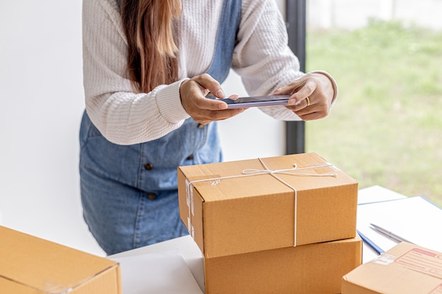 Una mujer que usa un teléfono inteligente para tomar fotografías frente a cajas de paquetería, cajas de paquetería para empacar mercancías, entregar mercancías a través de empresas de mensajería privadas. Conceptos de venta online y compra online.