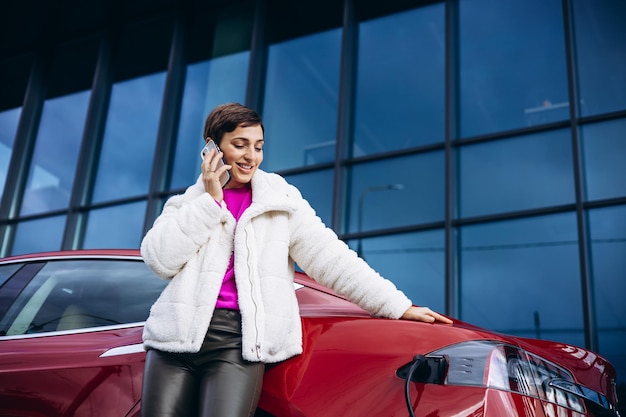 Mujer que usa el teléfono mientras carga un coche eléctrico junto al edificio de oficinas