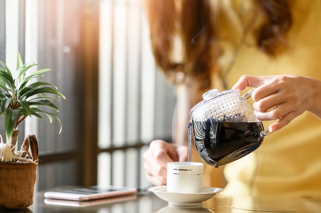 Mujer que vierte té caliente de la tetera de vidrio a la taza blanca mientras está sentado en el café