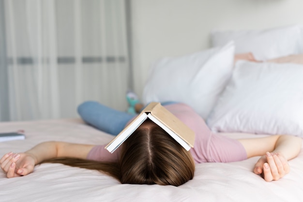 Foto mujer quedándose en la cama con un libro cubriéndose la cara