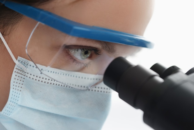 Mujer químico en gafas y máscara mirando a través del microscopio closeup desarrollo de vacuna
