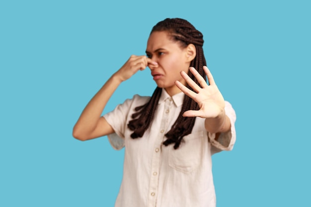 Mujer con rastas pellizcando la nariz deja de respirar mal olor disgustada por el olor a pedos su mueca expresando repulsión mostrando gesto de parada Estudio interior aislado en fondo azul
