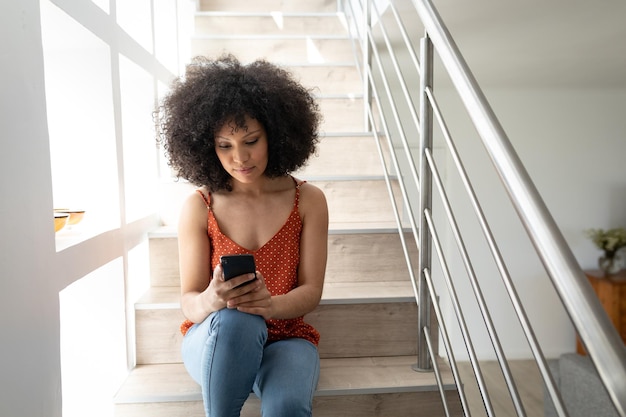 Mujer de raza mixta disfrutando del tiempo familiar en casa, distanciamiento social y autoaislamiento en cuarentena, sentada en las escaleras y usando un teléfono inteligente