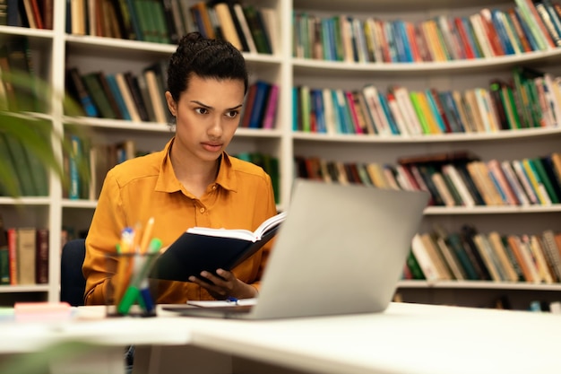 Mujer de raza mixta enfocada leyendo un libro y mirando una computadora portátil estudiando en línea sentada en la biblioteca gratis