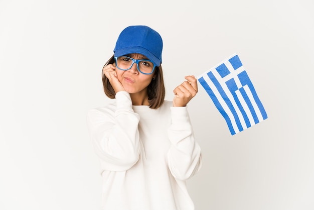 Mujer de raza mixta hispana joven sosteniendo una bandera de Grecia cubriendo los oídos con las manos.