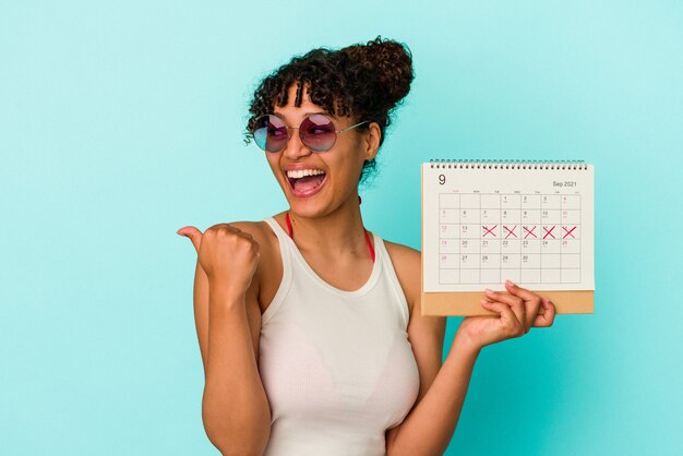 Mujer de raza mixta joven que sostiene el calendario aislado en la pared azul apunta con el dedo pulgar, riendo y despreocupado.