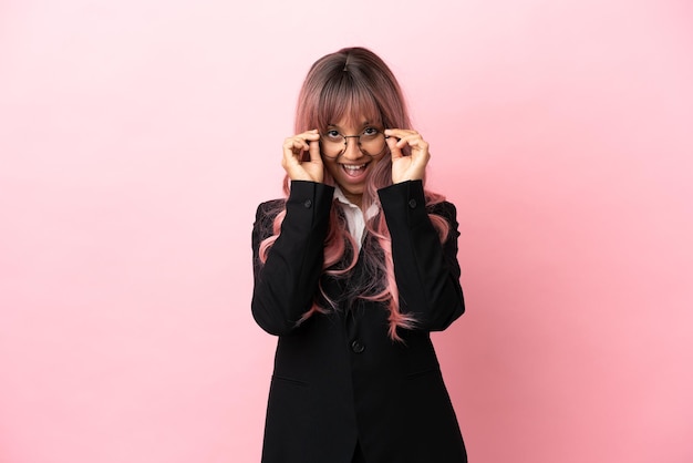 Mujer de raza mixta de negocios joven con cabello rosado aislado sobre fondo rosa con gafas y sorprendido