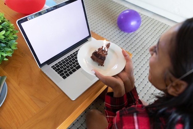 Mujer de raza mixta sosteniendo pastel de cumpleaños usando una computadora portátil para video chat. autoaislamiento durante la pandemia del coronavirus covid 19.