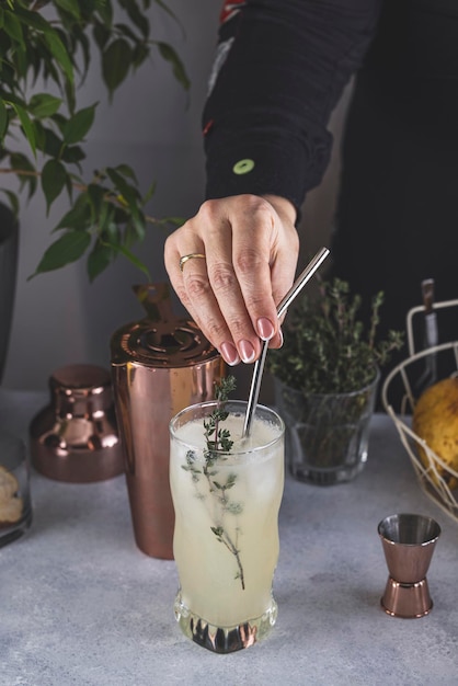 Mujer real preparando un cóctel Pear Collins en un vaso alto sobre una superficie de mesa gris claro rodeada de ingredientes y herramientas de bar