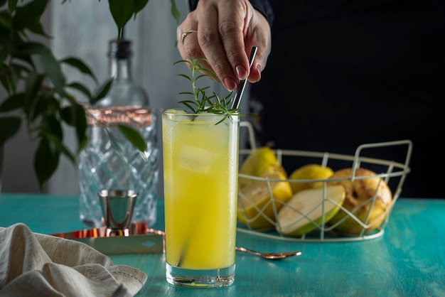 Mujer real preparando Pear Collins Cocktail en vaso alto o cócteles sin alcohol rodeados de ingredientes y herramientas de barra en la superficie de la mesa turquesa