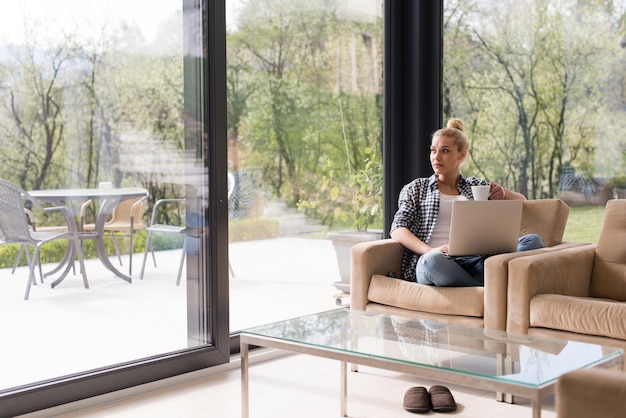 Mujer real que usa la computadora portátil en casa tomando café disfrutando de relajarse