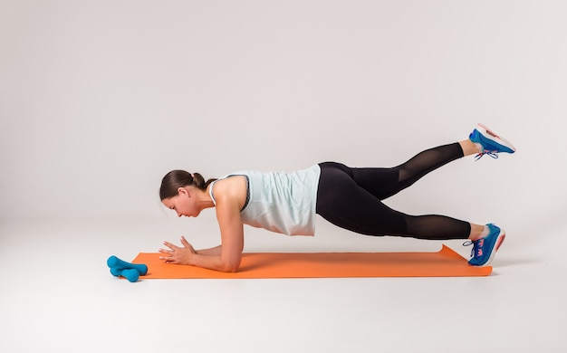 Una mujer realiza un ejercicio sobre una estera con la pierna levantada sobre un fondo blanco.