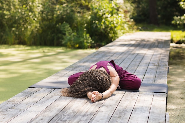 Mujer realizando ejercicio de balasana sentado en una colchoneta en el parque en una mañana soleada, postura del niño