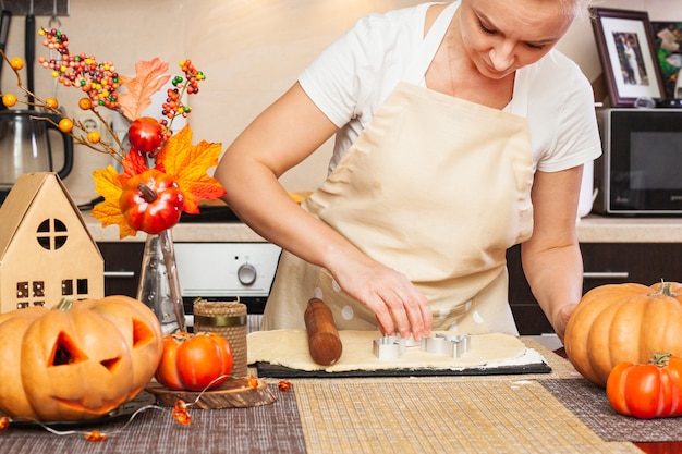 Una mujer recorta galletas para Halloween con un molde en la cocina con una decoración otoñal. Acogedora casa y preparación, Elaboración de galletas para Halloween.