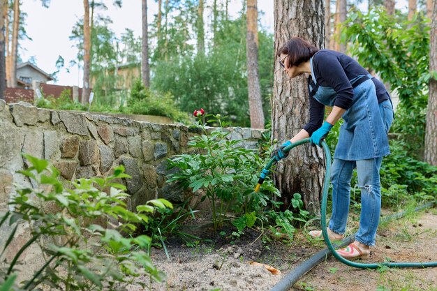 Mujer regando plantas en un lecho de flores en el patio trasero usando una manguera