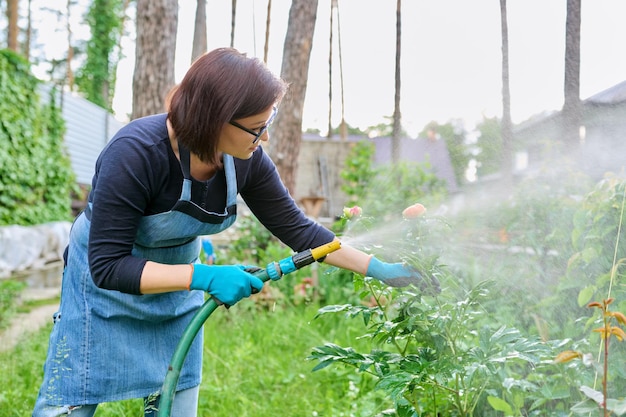 Mujer regando plantas en un lecho de flores en el patio trasero usando una manguera