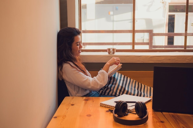 Mujer relajada en una cafetería leyendo un libro cerca de una ventana