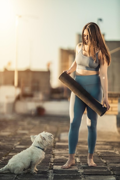 Una mujer relajada está preparando una alfombra de ejercicios para practicar yoga en una terraza en la azotea apoyada por su perro mascota al atardecer.