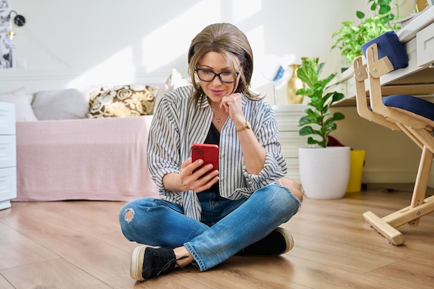 Mujer relajada de mediana edad sentada en el suelo en casa con un teléfono inteligente Mujer madura usando aplicaciones móviles para el trabajo, ocio, compras, negocios, comunicación, entretenimiento