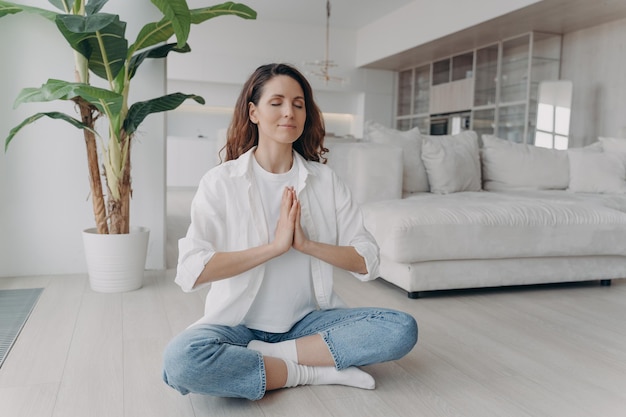 Una mujer relajada medita practicando yoga aliviando el estrés respirando profundamente sentada en el suelo en casa